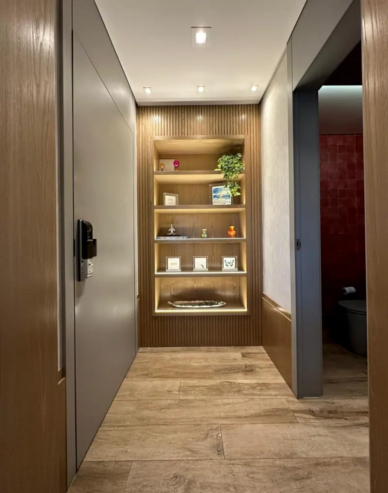 Corredor com painel de madeira, nicho iluminado com prateleiras e lavabo vermelho ao fundo