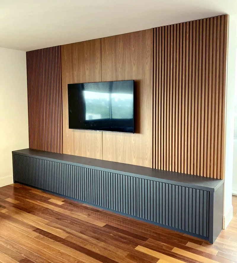 Painel de madeira ripada com TV na parede e rack baixo cinza escuro ripado, sobre piso de madeira